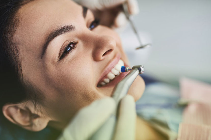 Close-up of a smiling woman receiving a fluoride treatment at a dental clinic, as the dentist polishes her teeth with a dental tool.