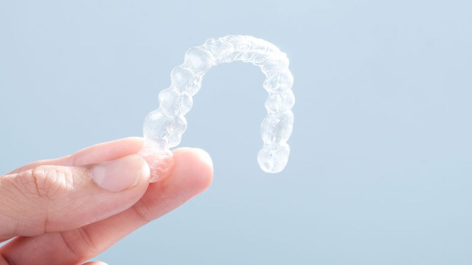 Close-up of hand holding a clear Invisalign aligner against a light blue pastel background.